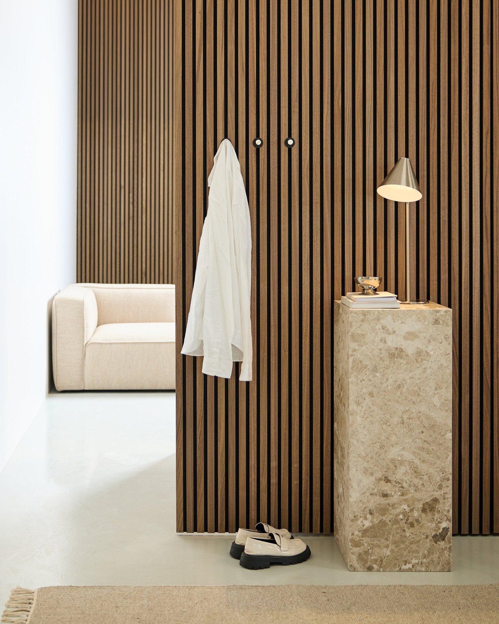Minimalistiskt rum med en nordisk akustikvägg i trälameller i Akupanel, vit skjorta på väggkrokar, piedestal i sten med lampa och böcker, beige soffa och vita skor på golvet.